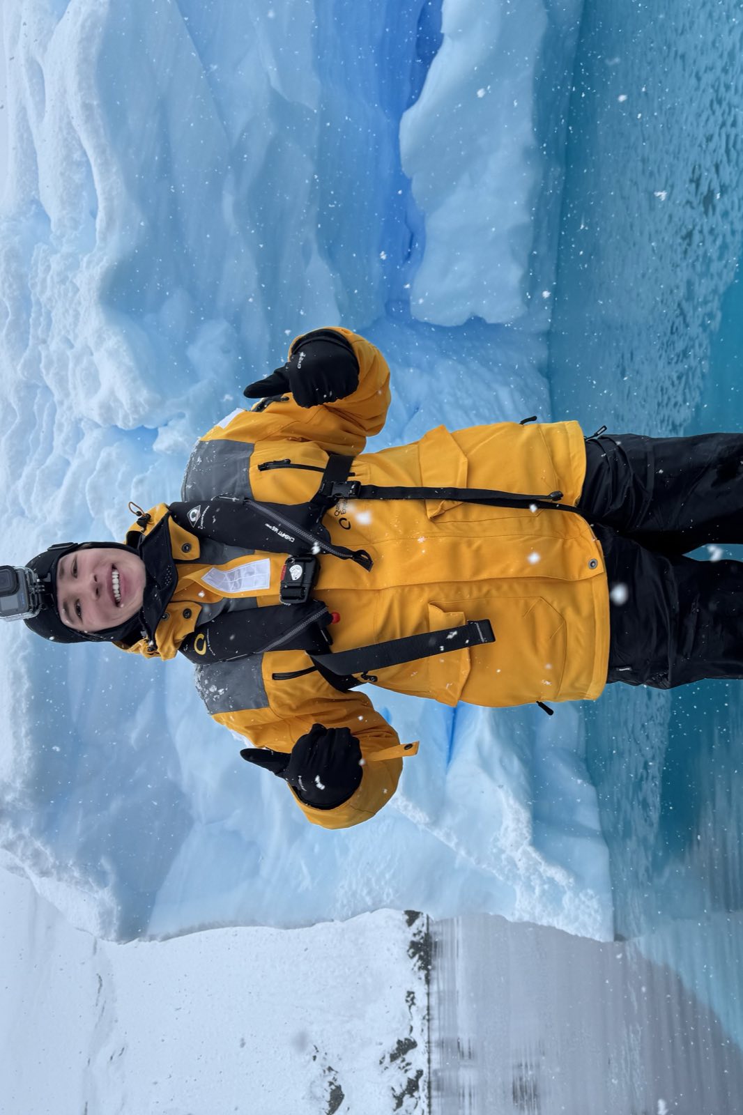John Jacobson in a yellow expedition parka and life vest, giving a thumbs up in front of a blue iceberg in Antarctica while snow falls.