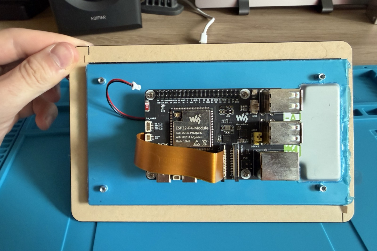 The ESP32-P4 development board mounted on the back of the seven inch touchscreen, held in one hand, with the ribbon cable joining the two.
